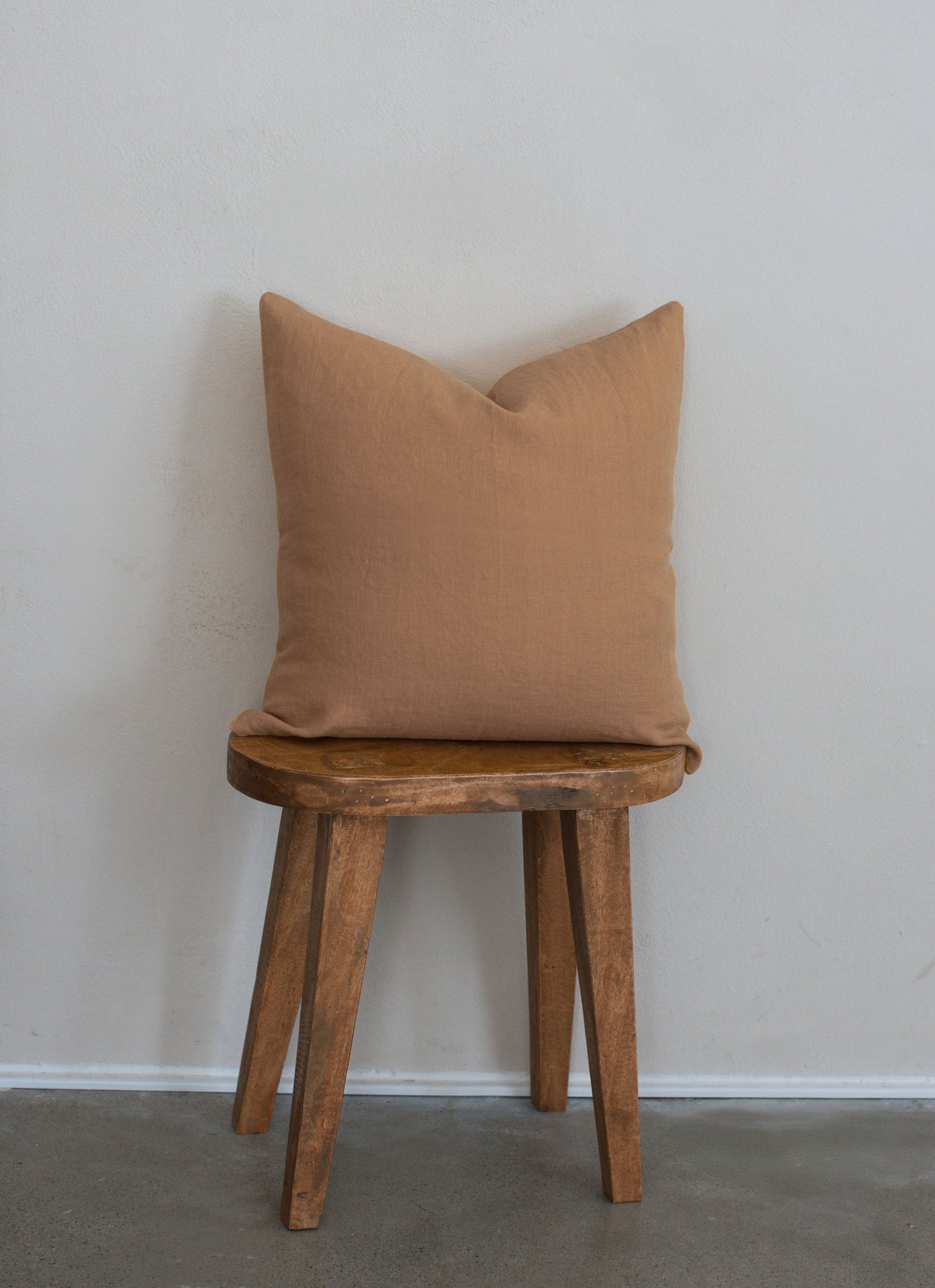 Tierra 100% linen accent pillow in beige displayed on a wooden stool against a white wall with a clean modern aesthetic.