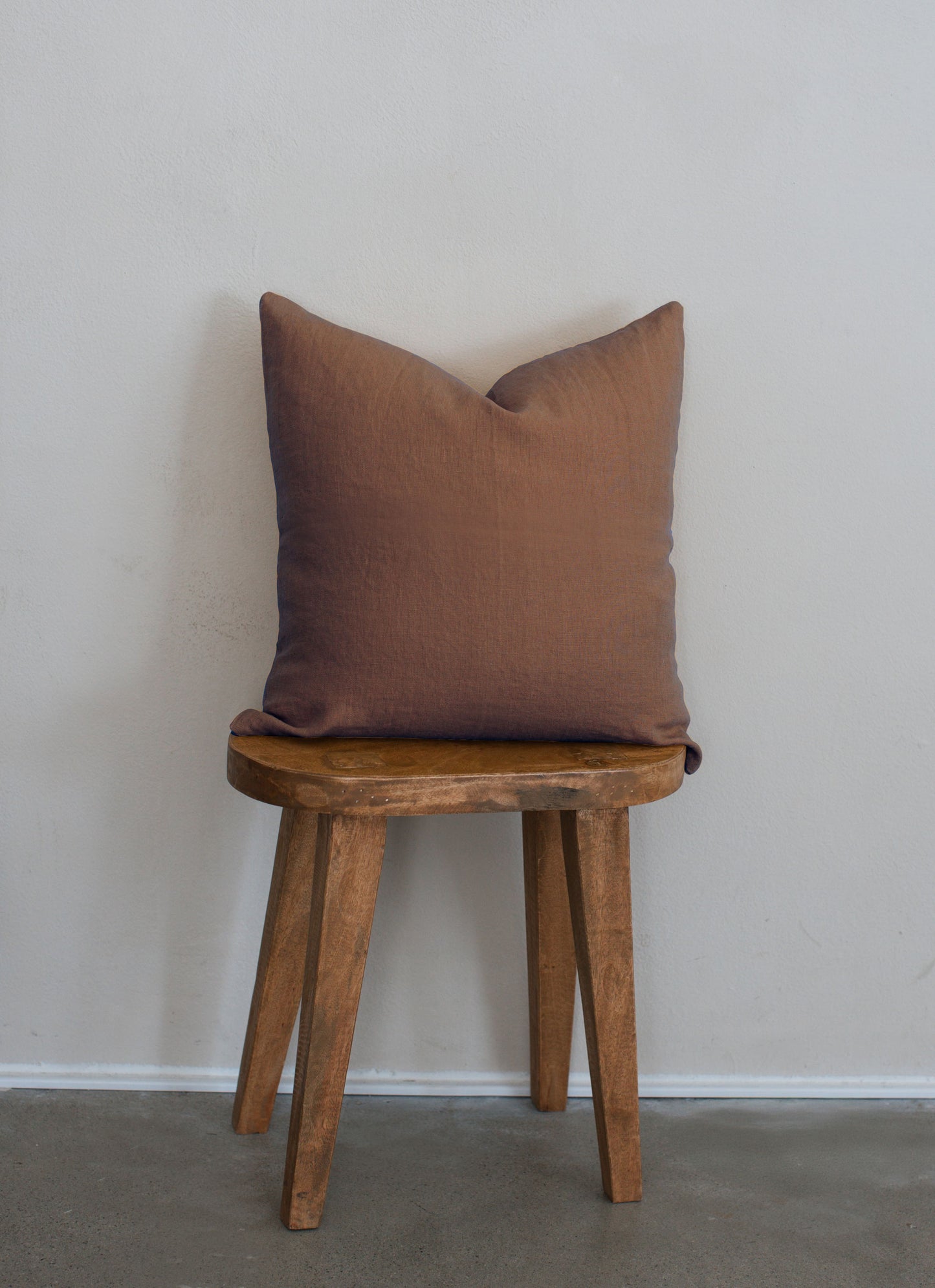 Tierra 100% linen accent pillow in brown displayed on a wooden stool against a white wall with a clean modern aesthetic.