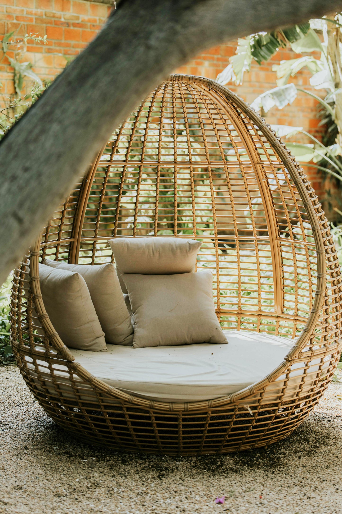 An outdoor lounge scene features Tierra’s 100% Linen Accent Pillow Cover styled inside a cozy wicker egg chair. Soft beige linen pillows are arranged on a circular cushioned seat, their natural texture and relaxed folds highlighted by dappled sunlight filtering through nearby trees. The woven rattan frame, surrounding greenery, and warm, earthy tones emphasize the breathable, temperature‑regulating qualities of pure linen. The image reflects Tierra Organic Spaces’ commitment to toxin‑free materials.