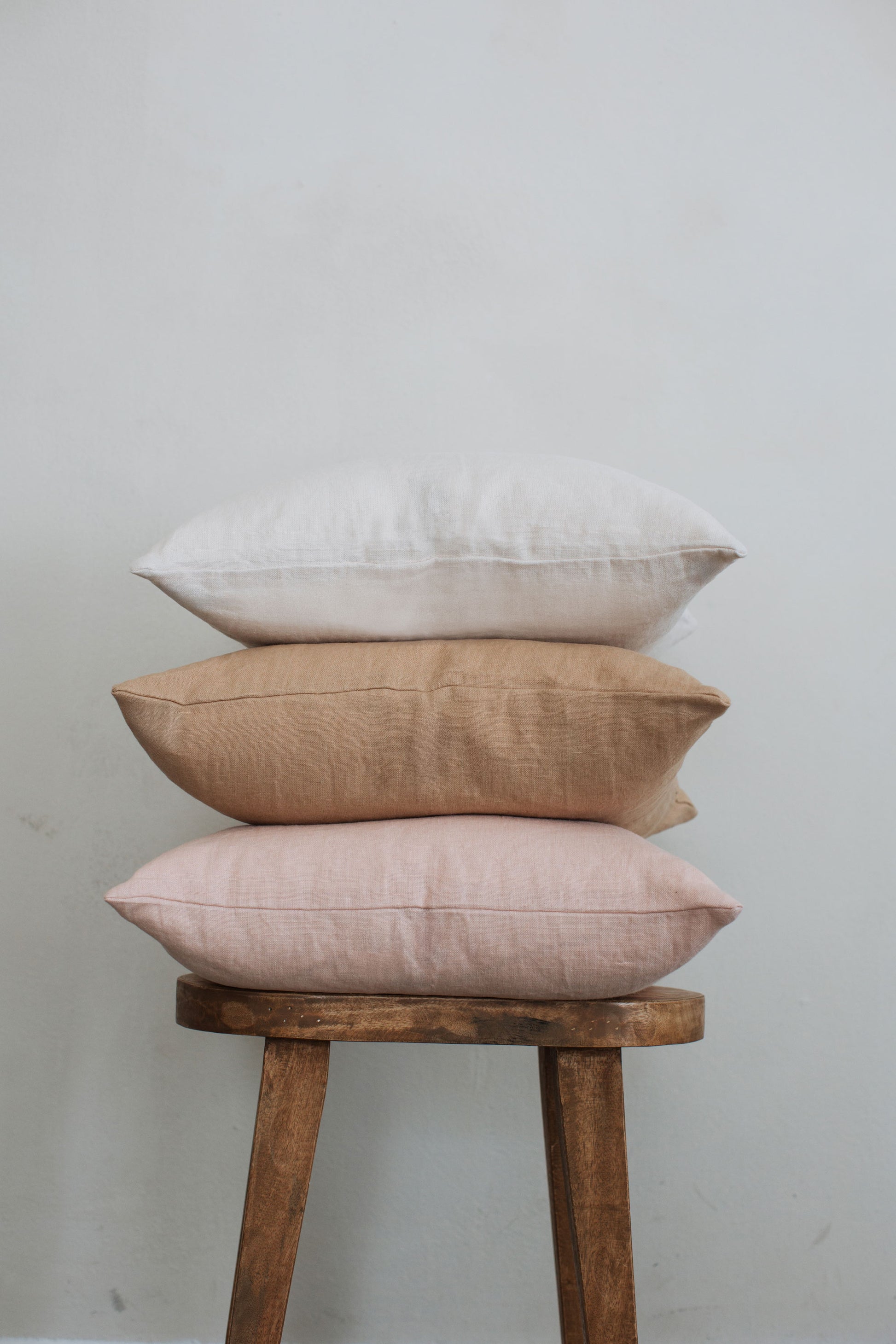Three neutral-colored Tierra 100% linen accent pillows stacked on a stool against a white wall showcasing the earth-toned linen color collection.