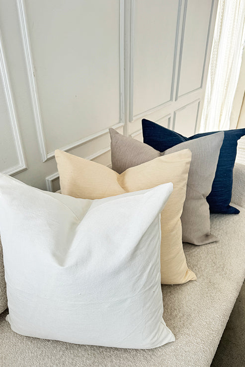 Tierra’s Organic Cotton Accent Pillow Cover styled in calm, neutral home settings. A beige sofa layered with large pillows in white, cream, beige, and deep blue, emphasizing the smooth texture and color variety of organic cotton.