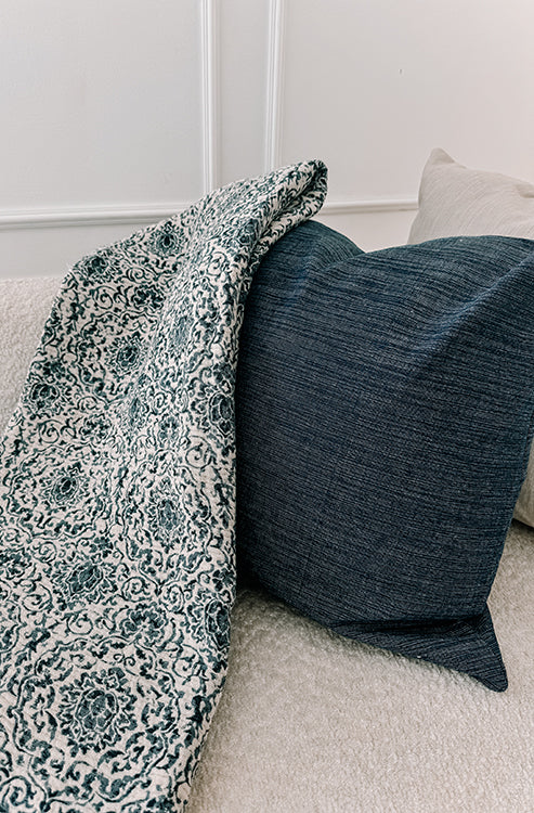 A close up of a denim blue textured pillow paired with a patterned throw, highlighting how the organic cotton accent pillow integrates seamlessly with layered textiles.