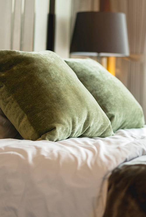 The image shows a neatly made bed with rich green velvet pillows beside a softly glowing lamp, emphasizing the warm, inviting feel of the fabric.