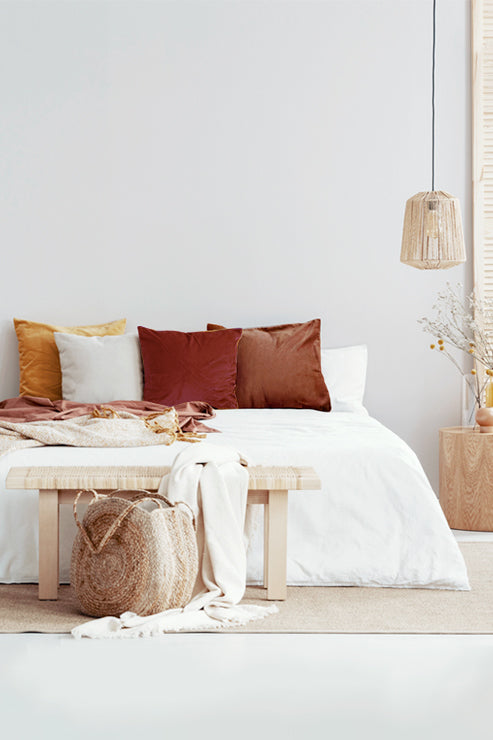 Boho-inspired bedroom with yellow, white and brown velvet pillows on a white bed, bright natural lighting, wood bench, and organic modern decor.
