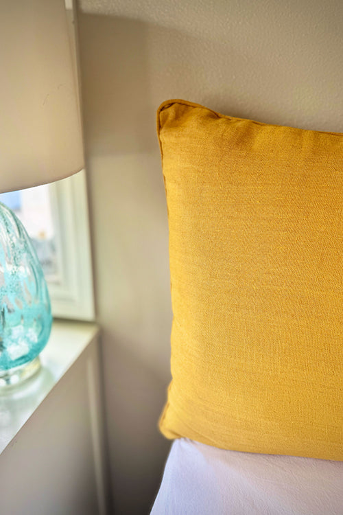 The image features a mustard‑toned pillow beside a softly glowing lamp, showcasing the warm elegance linen develops over time.