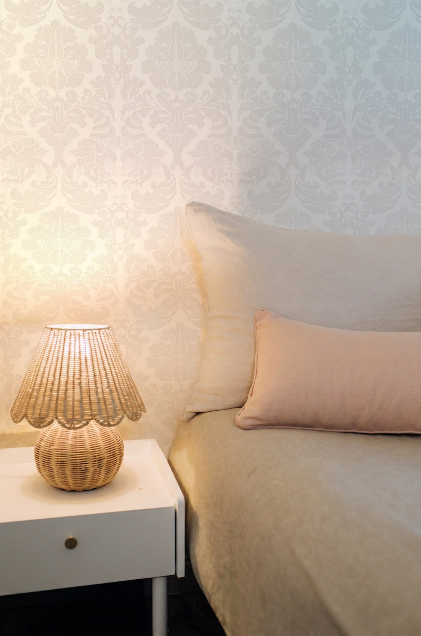 The image presents a cozy bedside vignette with a beige bedspread, pink pillow, patterned wallpaper, and a pleated lamp, underscoring the timeless, calming beauty linen brings to any space. 