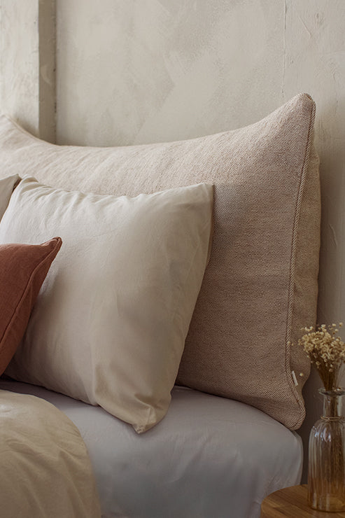 A softly lit bedroom features a large, textured headboard cushion in a warm neutral tone, styled behind neatly arranged pillows in beige and cream. The natural weave of Tierra’s Jute & Organic Cotton Blend Headboard Cushion Cover adds earthy strength and breathable comfort to the space, creating a calm, grounding backdrop for restorative rest.