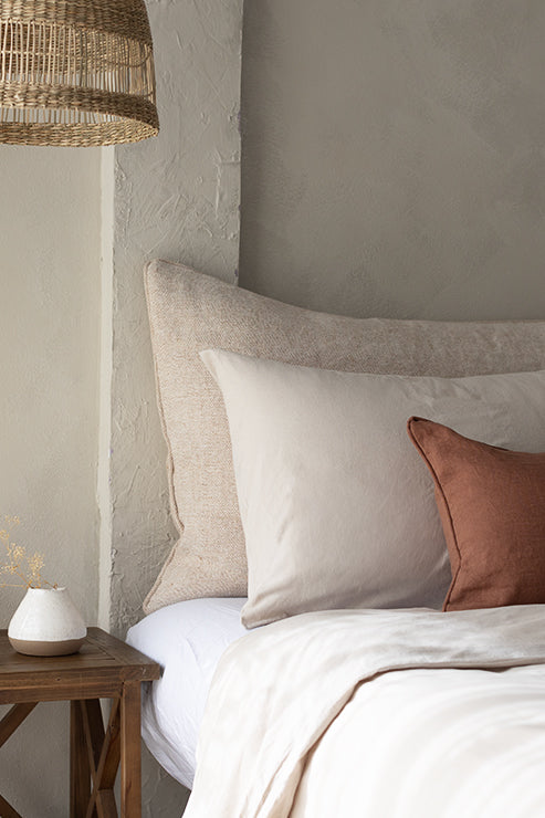 A cozy bedroom corner features a neatly made bed with neutral bedding and a large textured headboard cushion in warm beige tones. A small bedside table with a warm lamp and dried florals enhances the grounding, nature‑inspired aesthetic of Tierra’s Jute & Organic Cotton Blend Headboard Cushion Cover, reflecting its calming, breathable, and expertly crafted design.