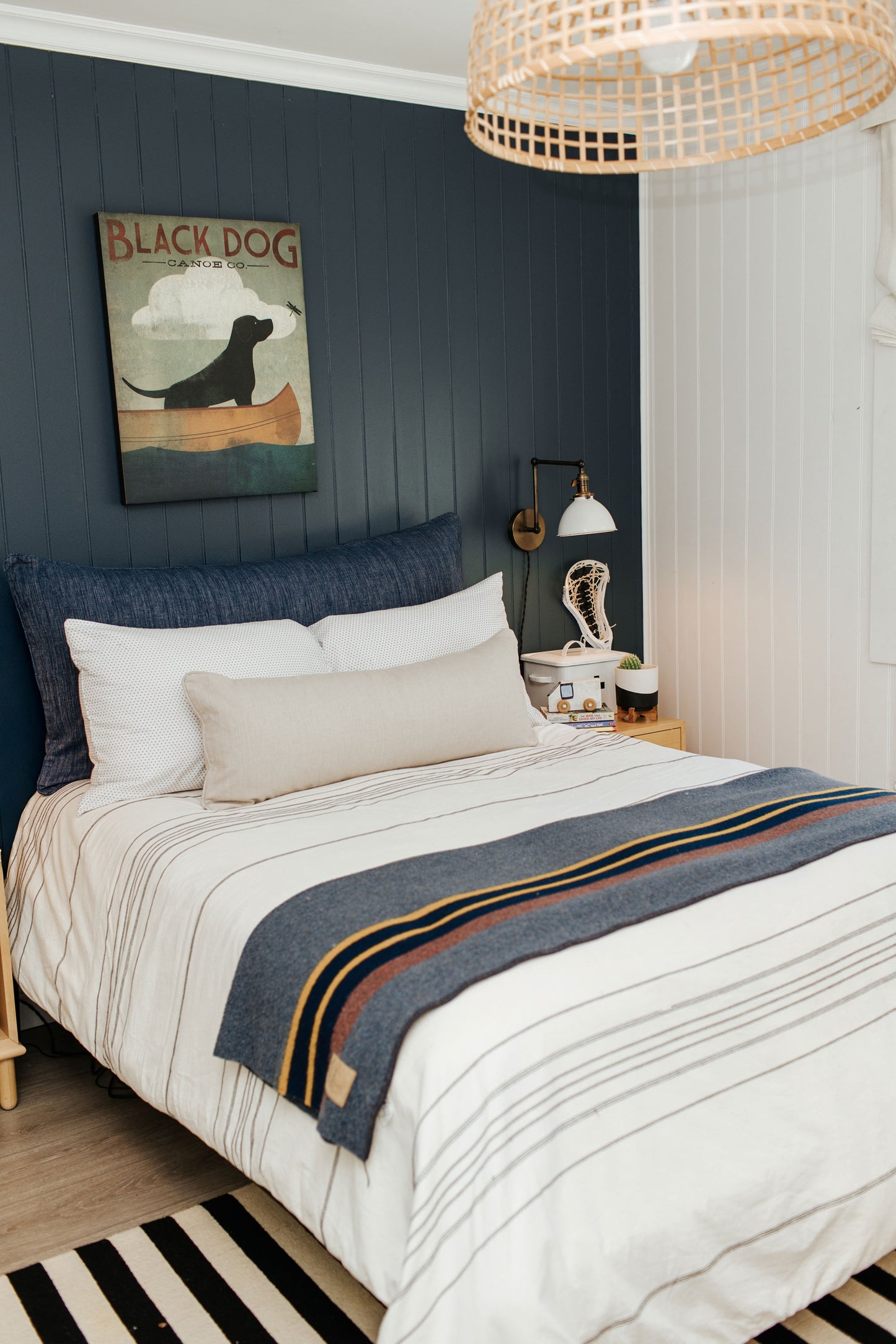 A neatly styled bedroom featuring a bed dressed in crisp white organic cotton bedding, Tierra's organic cotton Headboard Cushion in denim accented with a striped blanket in blue, orange, and gray tones. The clean, breathable textures highlight Tierra’s toxin‑free, 100% organic cotton craftsmanship. A navy accent wall and framed “BLACK DOG” artwork add understated character, while a bedside table with a lamp and small décor pieces reinforces the room’s simple, refined aesthetic.