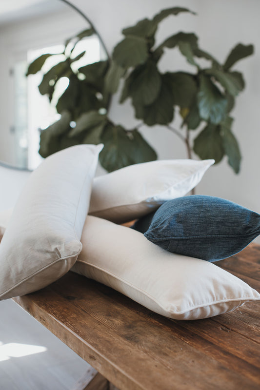 A stack of neutral-toned pillows arranged on a wooden table, showcasing soft, breathable organic cotton textures in white, beige, and deep navy. The pillows’ tailored design and smooth finishes reflect Tierra’s commitment to clean, hypoallergenic comfort and expert craftsmanship. A large green plant in the background adds natural warmth and complements the toxin‑free, OEKO‑TEX® certified materials.