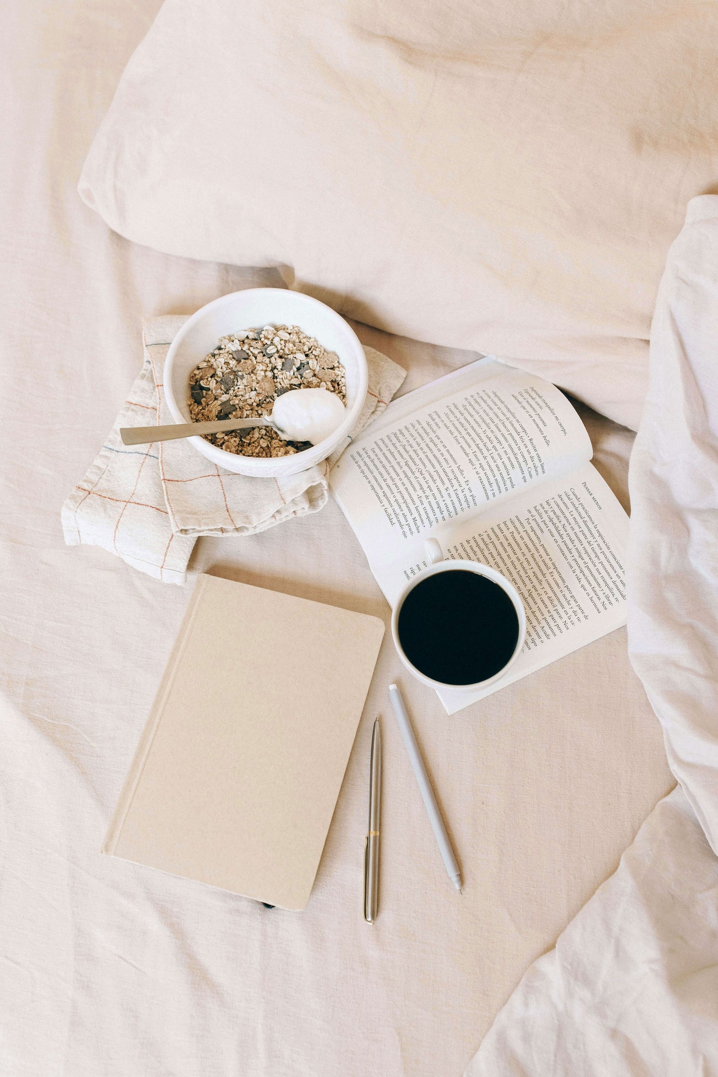 The image showcases Tierra’s Organic Cotton Bed Sheet through serene, neutral‑toned bedroom scenes. It features a cozy bed layered with soft beige organic cotton sheets, accompanied by a bowl of granola, an open book, a notebook, and a warm drink.  Evoking relaxed morning comfort.