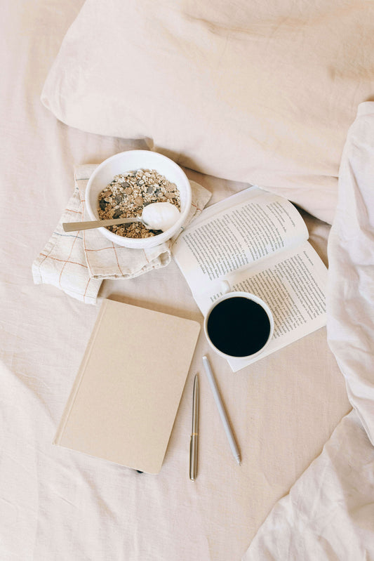 The image showcases Tierra’s Organic Cotton Bed Sheet through serene, neutral‑toned bedroom scenes. It features a cozy bed layered with soft beige organic cotton sheets, accompanied by a bowl of granola, an open book, a notebook, and a warm drink.  Evoking relaxed morning comfort.