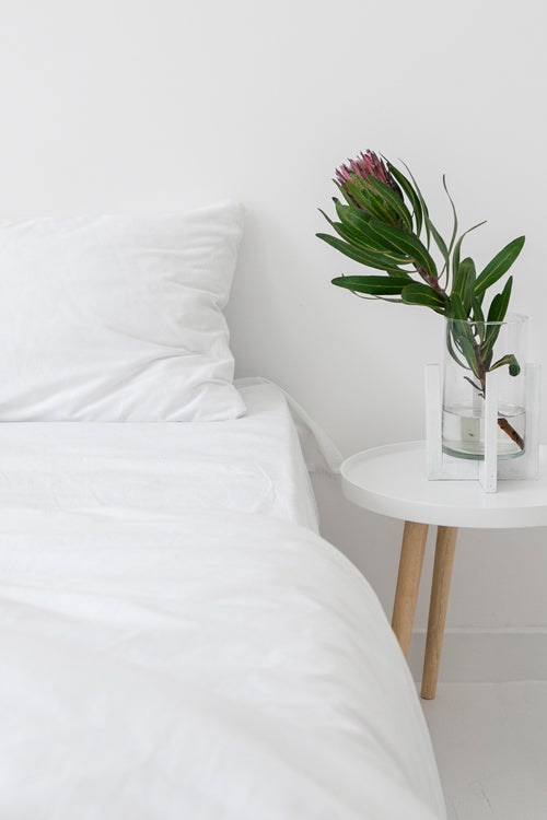 A neatly made bed dressed in crisp white bedding beside a minimalist round table holding a single protea flower, emphasizing the sheet’s refined simplicity and elevated design. 